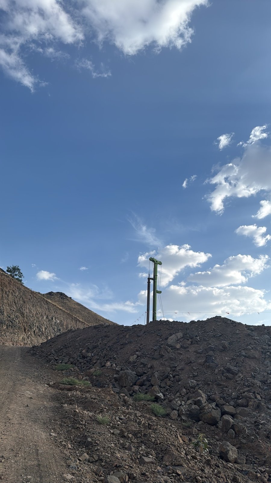A drilling rig mast visible above rock stockpiles beside an unpaved site route.