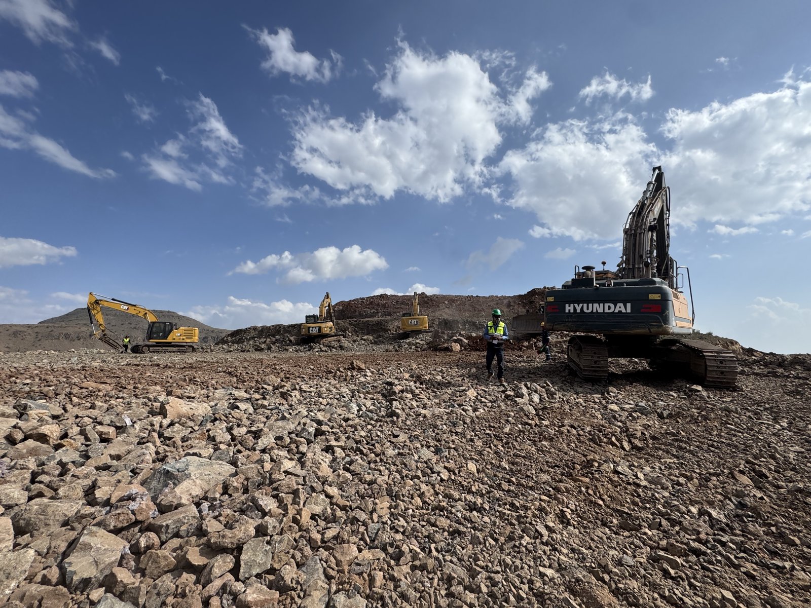 Tracked excavators and site personnel across a rocky earthworks area.