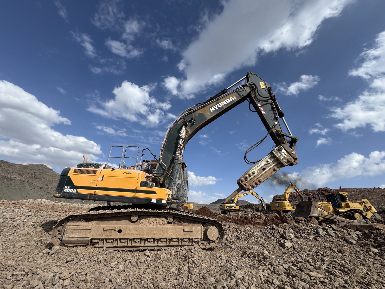 A Hyundai tracked excavator fitted with a hydraulic breaker, with earthmoving equipment across the rocky worksite.